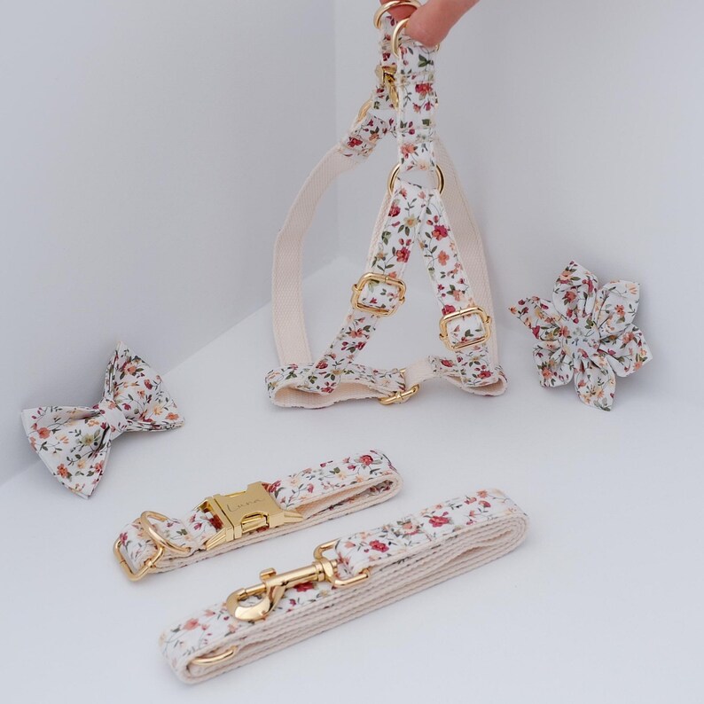 May include: A floral dog harness, leash, and bow tie set. The harness and leash are white with a floral pattern and gold hardware. The bow tie is a matching floral pattern.