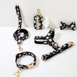 May include: A black and white daisy print dog collar, leash, harness, and bow tie with gold hardware. A small black and white daisy print bag with a gold zipper is also included.