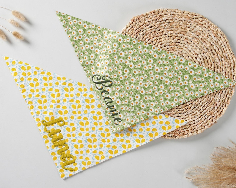 May include: Two triangular pet bandanas. One is white with yellow and blue floral pattern, embroidered with the name "Luna" in yellow. The other is green with white daisy pattern, embroidered with the name "Beanie" in black.