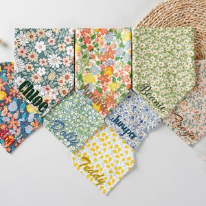 May include: A collection of personalized dog bandanas with floral patterns and names embroidered on them. The bandanas feature various designs, including daisies, lemons, and other colorful flowers. Names include Chloe, Aubree, Beanie, and others.
