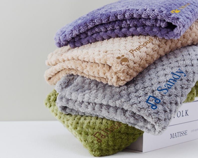 May include: Four plush pet blankets stacked on top of each other. The top blanket is purple, the second is beige, the third is gray, and the bottom is green. Each blanket has a different name embroidered on it: "Precious", "Sandy", "Lola", and "Matisse".