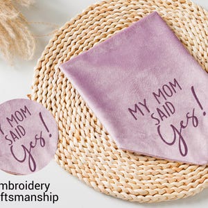 Puede incluir: Un pa&ntilde;uelo triangular de color lavanda con el texto "MY MOM SAID Yes!" en fuente cursiva. Tambi&eacute;n se ve un parche redondo a juego con el mismo texto. Los art&iacute;culos est&aacute;n sobre un salvamanteles circular tejido.