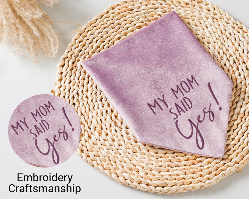 Puede incluir: Un pa&ntilde;uelo triangular de color lavanda con el texto "MY MOM SAID Yes!" en fuente cursiva. Tambi&eacute;n se ve un parche redondo a juego con el mismo texto. Los art&iacute;culos est&aacute;n sobre un salvamanteles circular tejido.