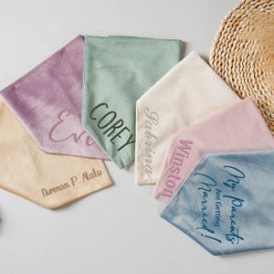 Puede incluir: Una colecci&oacute;n de pa&ntilde;uelos de tela suave personalizados en varios colores pastel, incluyendo lavanda, verde salvia y rosa p&aacute;lido. Cada pa&ntilde;uelo presenta texto bordado, con nombres y frases como "My Parents Are Getting Married!"