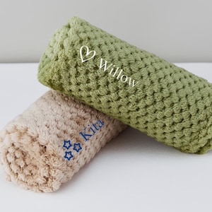 May include: Two plush, rolled-up blankets with embroidered names. The top blanket is green with the name "Willow" and a heart. The bottom blanket is beige with the name "Kita" and three stars.