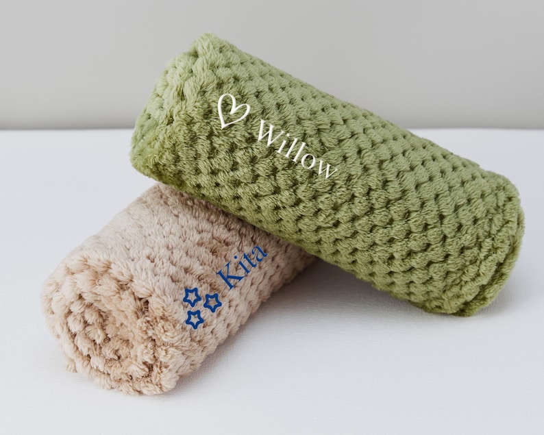 May include: Two plush, rolled-up blankets with embroidered names. The top blanket is green with the name "Willow" and a heart. The bottom blanket is beige with the name "Kita" and three stars.
