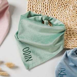 Puede incluir: Primer plano de tres pa&ntilde;uelos de terciopelo en colores pastel: rosa, verde menta y azul. El pa&ntilde;uelo verde menta tiene bordado el nombre "ODIN". Los pa&ntilde;uelos se muestran sobre una superficie blanca con una cesta tejida en el fondo.
