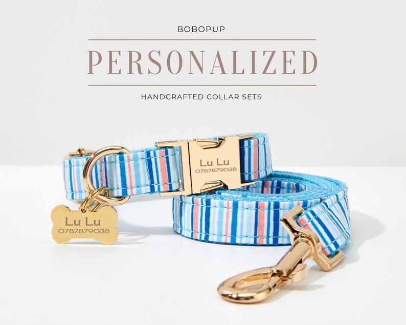 May include: Personalized dog collar and leash set with blue, white, and coral stripes. The gold-colored buckle and tag are engraved with the name "Lu Lu" and a phone number. The text "BOBOPUP" and "PERSONALIZED HANDCRAFTED COLLAR SETS" are displayed.