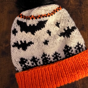 May include: A knitted Halloween beanie in cream, orange, and black. The hat features a black pom-pom, orange brim, and a pattern of black bats and stars. The top of the hat has an orange and black zigzag design.