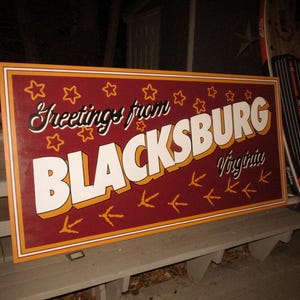 May include: A red and gold sign with white lettering that reads "Greetings from Blacksburg, Virginia". The sign has a border of gold stars and a pattern of white footprints.