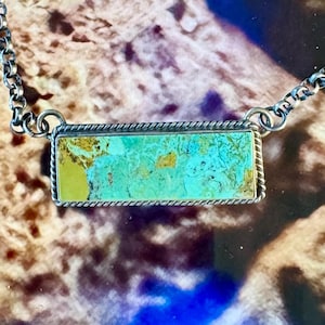 May include: A rectangular turquoise stone pendant set in a silver frame with a twisted rope border. The stone features a mix of vibrant teal, blue, and earthy yellow-brown matrix patterns. It is attached to a silver rolo chain necklace.