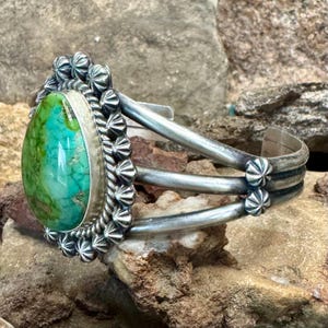 May include: A silver cuff bracelet featuring a large, pear-shaped turquoise stone with green and blue marbling. The stone is surrounded by a decorative scalloped silver border and a twisted wire frame. The band consists of three parallel silver wires.