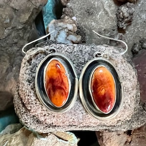 May include: A pair of silver earrings with orange and red agate stones set in oval bezels. The earrings have a simple wire hook closure.
