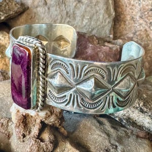 May include: A silver cuff bracelet featuring a rectangular, deep purple stone set in a twisted metal border. The band is decorated with embossed geometric diamond shapes and curved line patterns, creating a textured, metallic appearance.