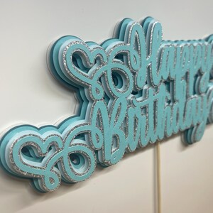 May include: A layered, light blue "Happy Birthday" cake topper with silver glitter accents. The script lettering is multi-dimensional, with a wooden stick for insertion into a cake. The topper is set against a white background.