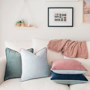 May include: A white couch with four throw pillows. The pillows are teal green, light blue, pink, and navy blue. The pillows have a light pink, dark blue, and light green trim. A pink knitted throw is draped over the back of the couch.
