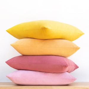 May include: A stack of four decorative throw pillows in shades of yellow, orange, pink, and light pink. The pillows are arranged in a pyramid shape, with the yellow pillow on top and the light pink pillow on the bottom.