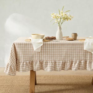 May include: A beige and white gingham tablecloth with a ruffled edge draped over a table. On the table sits a white ceramic vase containing white flowers, a beige ceramic bowl, a white cloth napkin, and a brown ceramic mug.