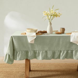 May include: A sage green tablecloth with a ruffled edge covers a wooden table. A white vase with yellow and white flowers sits on the table, along with a bowl, napkins, and a mug. The table is set on a woven rug.