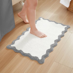 May include: A white bath mat with a gray scalloped border. The mat is made of soft, absorbent material and is perfect for drying your feet after a shower.