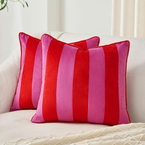 May include: Two square decorative pillows with a bold striped pattern in red and pink. The pillows are placed on a white sofa, offering a pop of color and a modern aesthetic. The pillows have a soft, velvety texture.