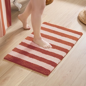 May include: A rectangular bath mat with alternating stripes of orange and white. The mat is made of a soft, absorbent material, and is placed on a light-colored wooden floor. The mat is designed to absorb water and prevent slips.