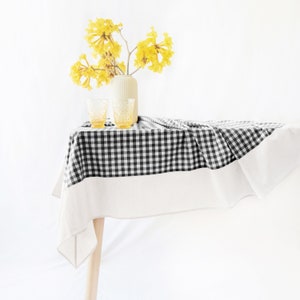May include: A black and white checkered tablecloth with a white border, draped over a table with a wooden leg. Two clear glass tumblers and a vase with yellow flowers are on the table.