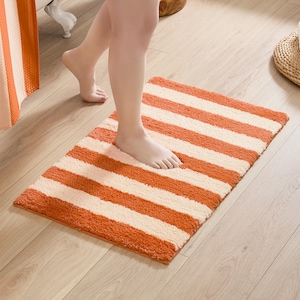 May include: Orange and cream striped bath mat. The rectangular mat features thick, plush stripes of orange and cream. The mat is placed on a light wood floor. A person's foot is on the mat.