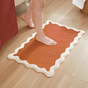 May include: A terracotta bath mat with a white scalloped border. The mat is on a light wood floor.