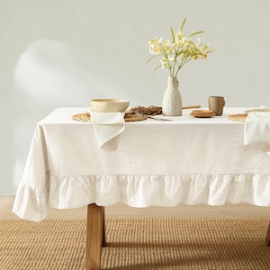 May include: A white linen tablecloth with a ruffled edge covers a wooden table. The table is set with a bowl, a vase of flowers, and a mug. The tablecloth is on a woven rug.