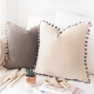May include: Two decorative throw pillows with a pom-pom trim. The pillow on the left is gray and the pillow on the right is beige.