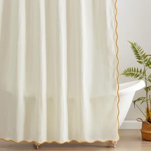 May include: Cream-colored waffle-weave shower curtain with a scalloped edge in a golden yellow hue. The curtain hangs in front of a white bathtub, with a fern and a woven basket in the background. The curtain adds a touch of elegance to any bathroom.