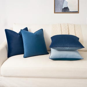 May include: Four square throw pillows in varying shades of blue velvet. Two pillows are upright, one in deep navy and one in a medium blue. Two pillows are stacked horizontally, with a dark navy top pillow and a light blue bottom pillow.