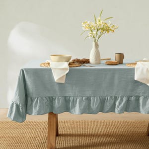 May include: A light blue tablecloth with a ruffled edge covers a wooden table. A vase of white flowers, a bowl, and a mug are on the table. The table is set on a woven jute rug.