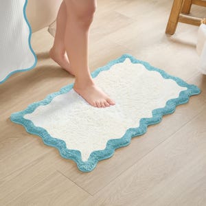 May include: A rectangular, off-white bath mat with a scalloped, light blue border. The mat is made of a soft, absorbent material and is placed on a light wood floor. The mat is designed to be placed in front of a bathtub or shower.