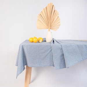 May include: A blue and white striped tablecloth draped over a wooden table. A white vase holds a large, dried palm leaf. Three lemons are arranged on the table. The scene is set against a white background.