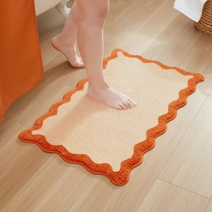 May include: A beige bath mat with an orange scalloped border. The mat is made of soft, plush material and is designed to absorb water.