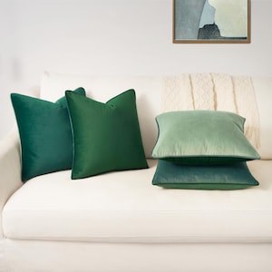 May include: Four square throw pillows in varying shades of green velvet. Two pillows are a deep forest green, while the other two are a lighter sage green. The pillows have a smooth, soft texture and are arranged in a stack and side-by-side.