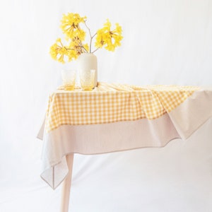 May include: A yellow and white checkered tablecloth with a beige border is draped over a wooden table. Two yellow-tinted glasses and a white vase with yellow flowers are on the table. The background is white.