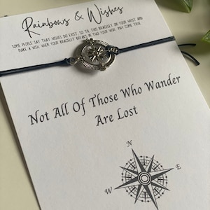 May include: A black string bracelet with a silver compass charm. The bracelet has a card attached to it that reads "Rainbows & Wishes" and "Not All Of Those Who Wander Are Lost".