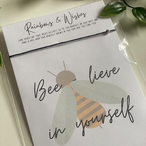 Op de afbeelding: Een witte kaart met de tekst "Rainbows & Wishes" en "Bee-lieve in yourself" met een cartoon-bij-illustratie. De kaart is bevestigd aan een zwart koordarmbandje met een kleine grijze kraal.