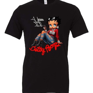 May include: Black t-shirt featuring a Betty Boop graphic. The cartoon character is depicted in a red top and distressed jeans, holding a phone. The words "Betty Boop" are in red script, with "Urban BB" in the upper left.