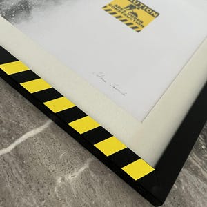 May include: A framed print featuring a black and white image with a yellow and black caution sign that reads CAUTION AREA UNDER CONSTRUCTION. The frame is black with yellow and black diagonal hazard stripes along the edges and a white mat.