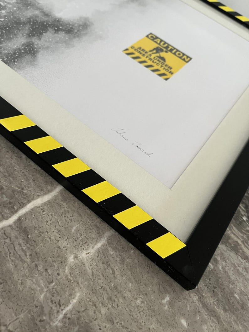 May include: A framed print featuring a black and white image with a yellow and black caution sign that reads CAUTION AREA UNDER CONSTRUCTION. The frame is black with yellow and black diagonal hazard stripes along the edges and a white mat.