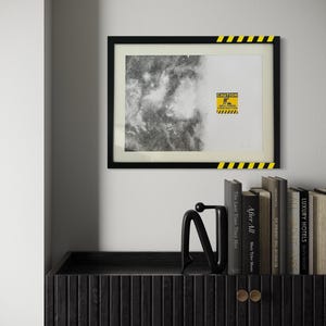 May include: A framed rectangular print featuring a black and white nebula image on the left and a yellow caution sign on the right with the text CAUTION AREA UNDER CONSTRUCTION. The black frame has yellow and black hazard stripes in two corners.