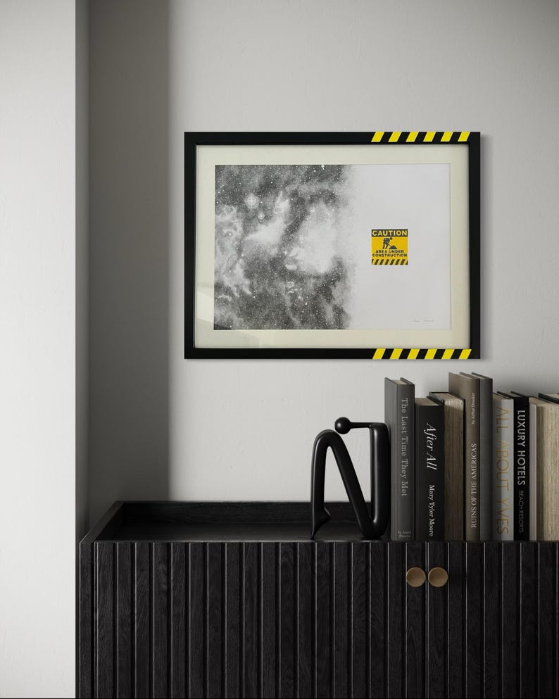 May include: A framed rectangular print featuring a black and white nebula image on the left and a yellow caution sign on the right with the text CAUTION AREA UNDER CONSTRUCTION. The black frame has yellow and black hazard stripes in two corners.