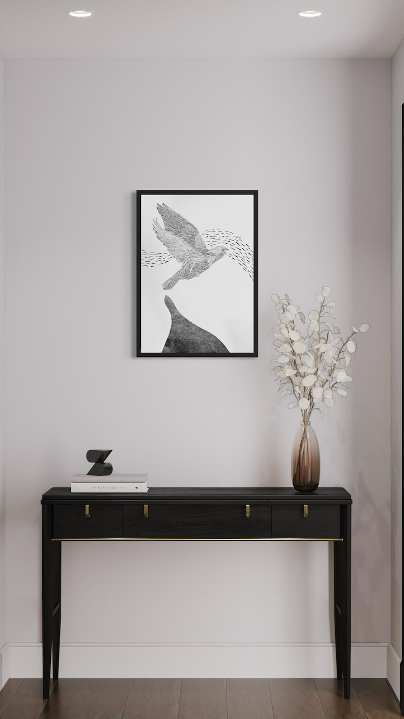 K&ouml;nnte beinhalten: Ein schwarz gerahmter Kunstdruck mit einer detaillierten schwarz-wei&szlig;en Illustration eines fliegenden Vogels &uuml;ber einem dunklen Berggipfel, platziert &uuml;ber einem schwarzen Konsolentisch mit goldenen Beschl&auml;gen, einer Glasvase und B&uuml;chern.