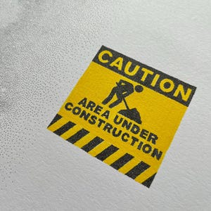 May include: A square yellow sticker featuring a black border and black text that reads CAUTION AREA UNDER CONSTRUCTION. The design includes a black graphic icon of a person digging with a shovel and a black and yellow striped pattern at the bottom.