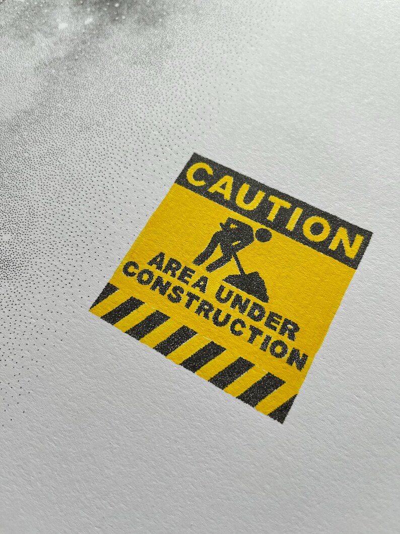 May include: A square yellow sticker featuring a black border and black text that reads CAUTION AREA UNDER CONSTRUCTION. The design includes a black graphic icon of a person digging with a shovel and a black and yellow striped pattern at the bottom.
