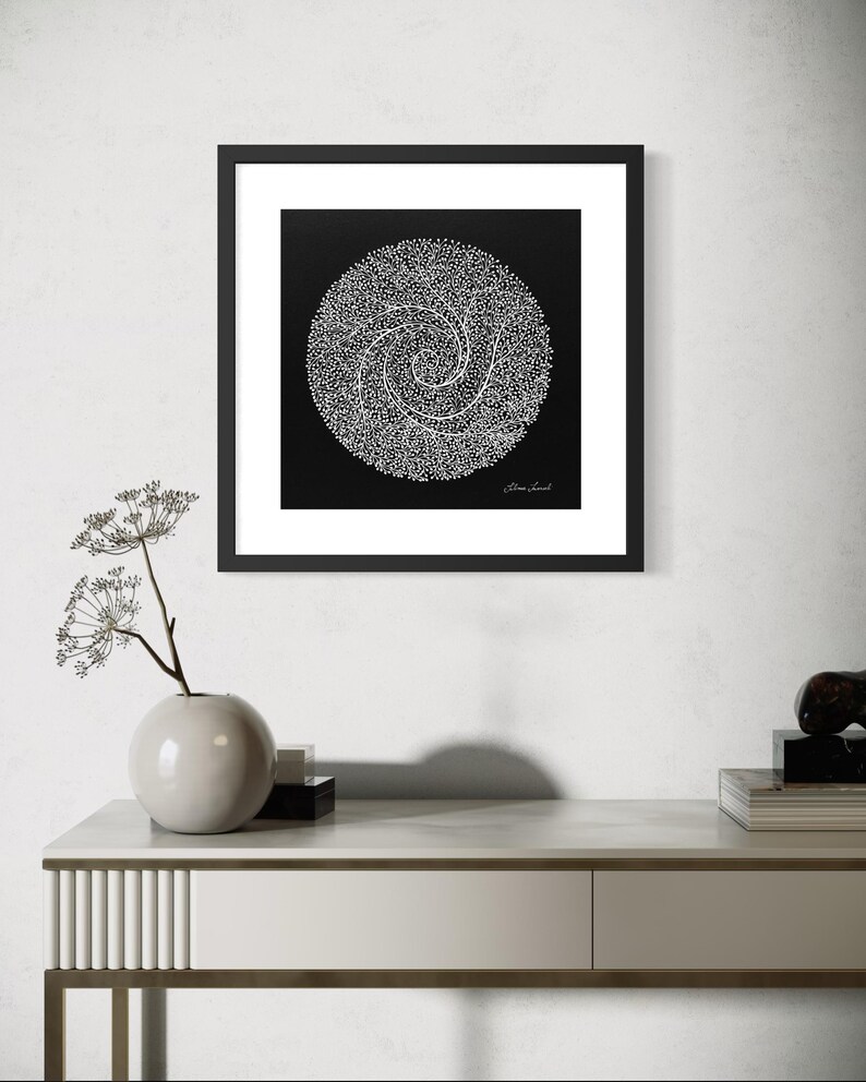 May include: A square framed wall art piece featuring a black background with a white circular design composed of intricate, swirling dotted lines. The artwork includes a signature in the bottom right corner and is enclosed in a thin black frame.